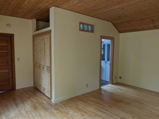 An inside view at a living space. The walls are painted a bright off-white, and there is wood paneling on the floor and ceiling. At the middle is the entry to another room.