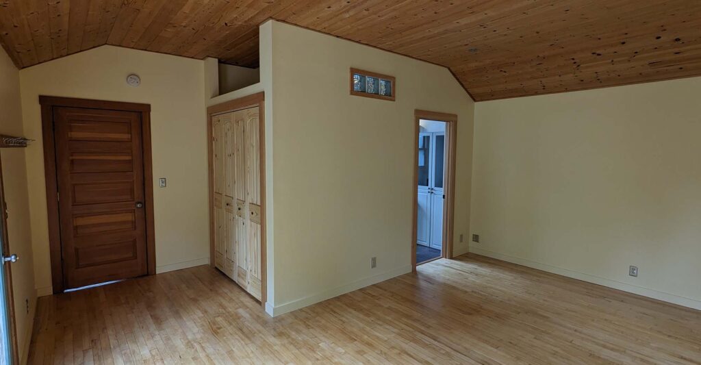 An inside view at a living space. The walls are painted a bright off-white, and there is wood paneling on the floor and ceiling. At the middle is the entry to another room.