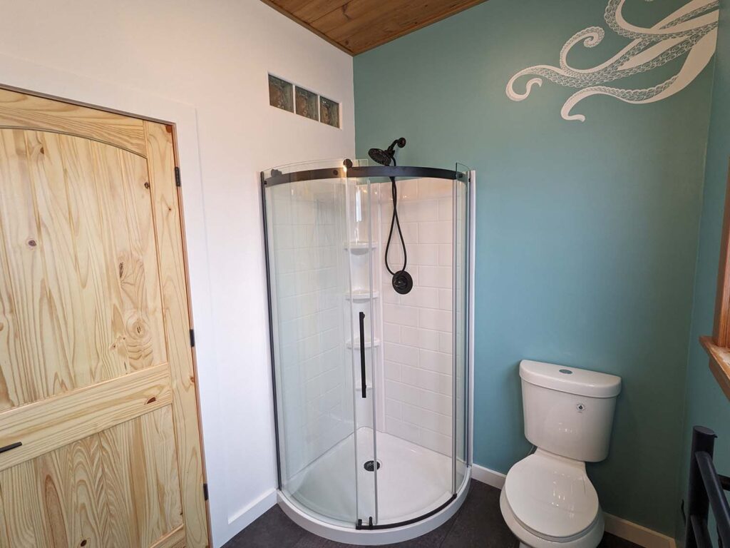 An interior view of a bathroom. The walls are painted teal, and one of them has an octopus feature painted on it. There is a small corner shower next to a beautiful wood door.