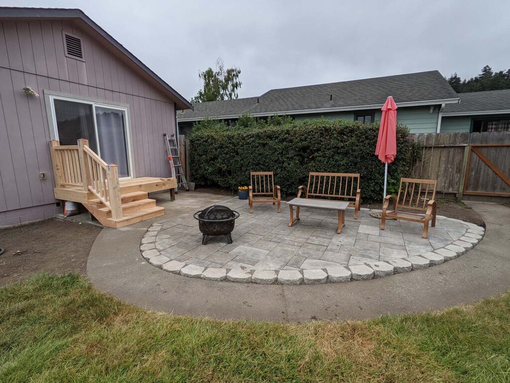 A back yard lounge area with newly-installed cement bricks, new landscaping, a fire bowl, and some newly-built stairs.