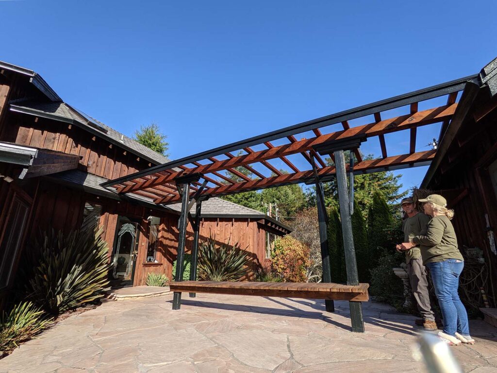 Two people stand looking up at a beautiful glass and stained wood pergola